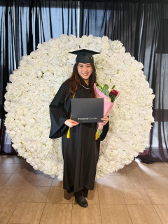 Student in graduation gown and cap standing in front of a decorative flower wall